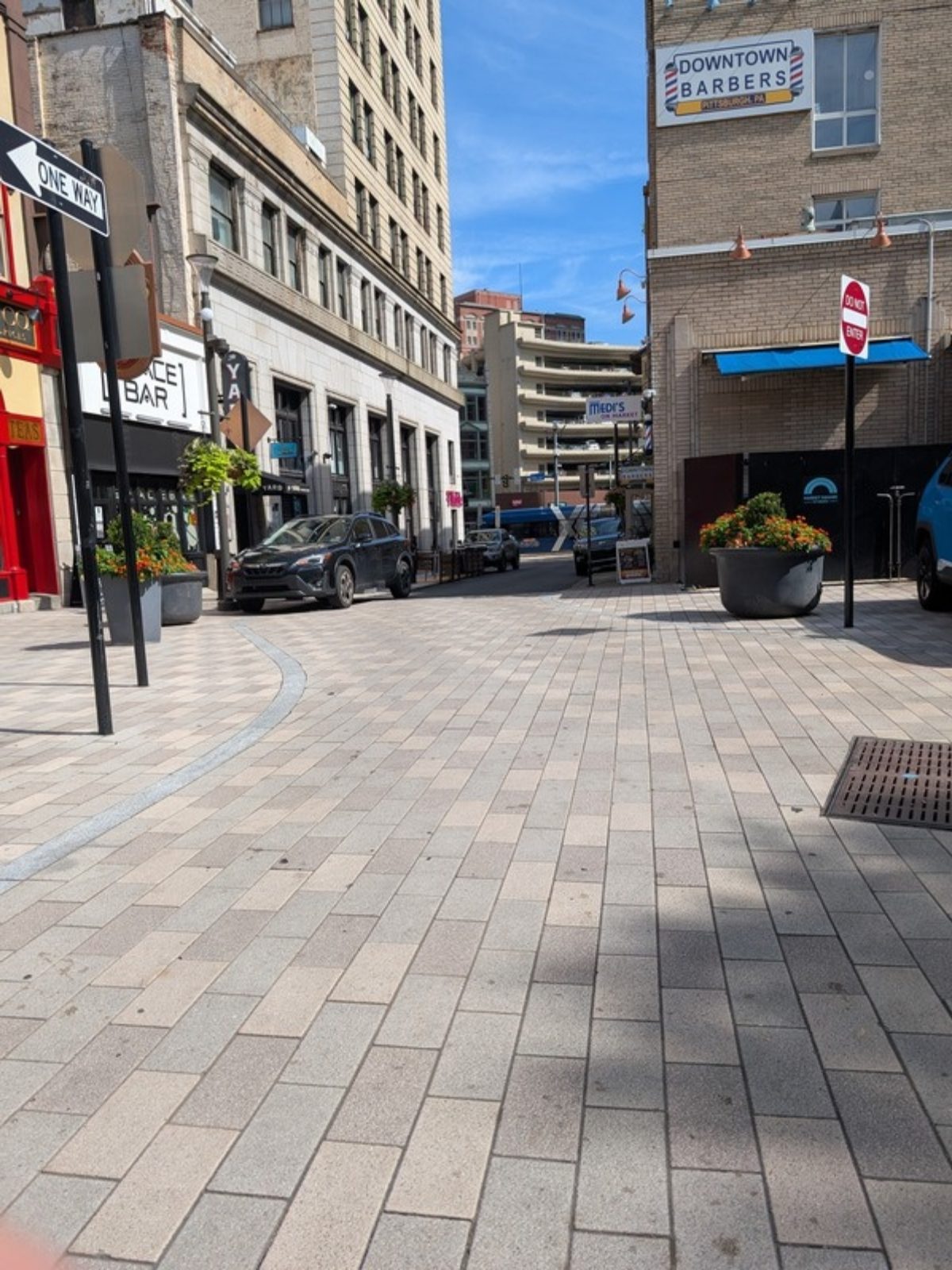 Looking out of Market Square toward a side street, at seated height. Light grey paving stones run flat in the foreground and continue into the street without a curb, with a curved band of darker stone along the left. A dark SUV is parked on the left side of the street and a parking garage stands a block away. A large planter of yellow flowers, a do not enter sign, and a metal drain grate sit at the right edge of the paving.