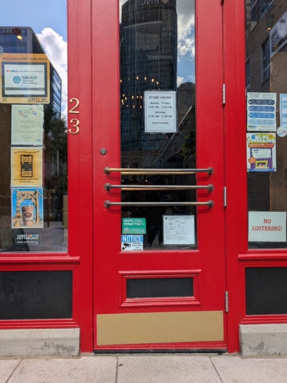 Close view of the red entrance door from directly in front, at seated height. A tall glass panel runs down the middle of the door with three horizontal brass pull bars mounted across it, one above another. A brass kick plate covers the bottom of the door. Paper signs showing store hours and permits are taped to the glass, and the number 23 is on the frame to the left. The paving outside meets the base of the door with no step.