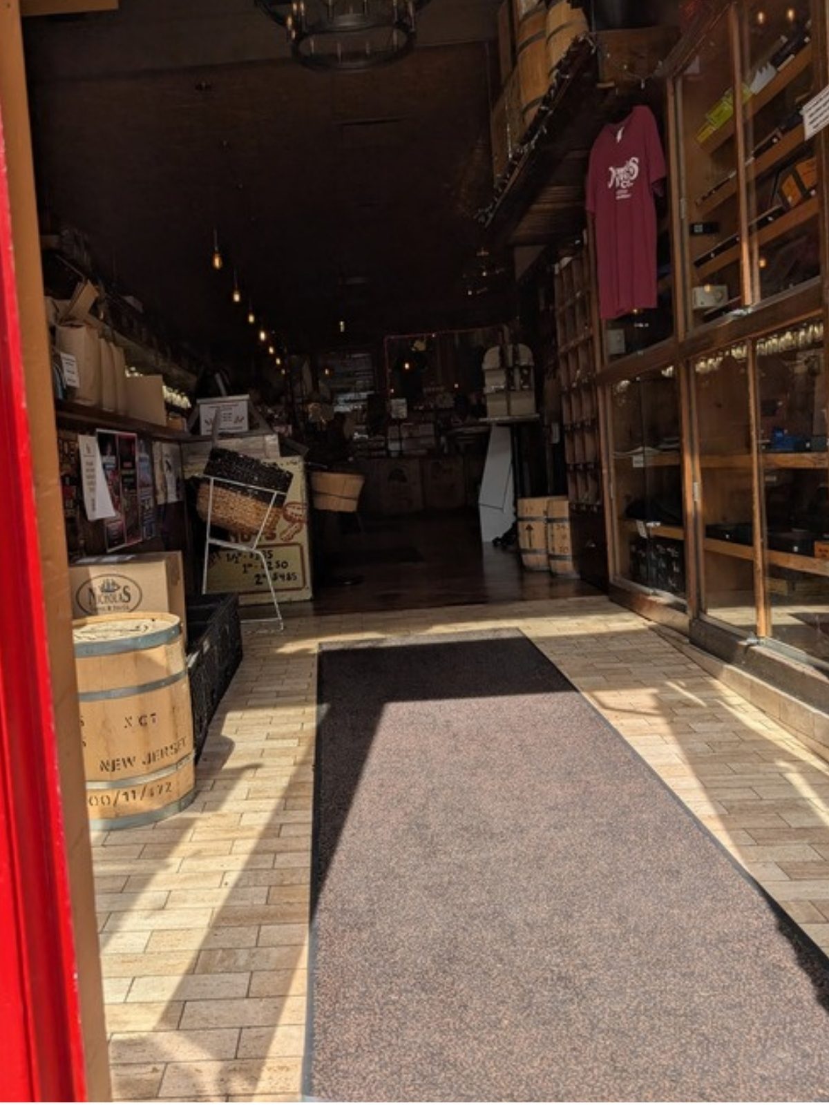 View from the open doorway looking straight into the shop at seated height. The red door frame fills the left edge. Just inside, a pale tiled entry floor runs forward with a long dark rubber-edged mat laid down the middle of it. Wooden barrels and boxes of coffee stand against the left wall and glass display cabinets line the right wall. The shop runs back into darker space.