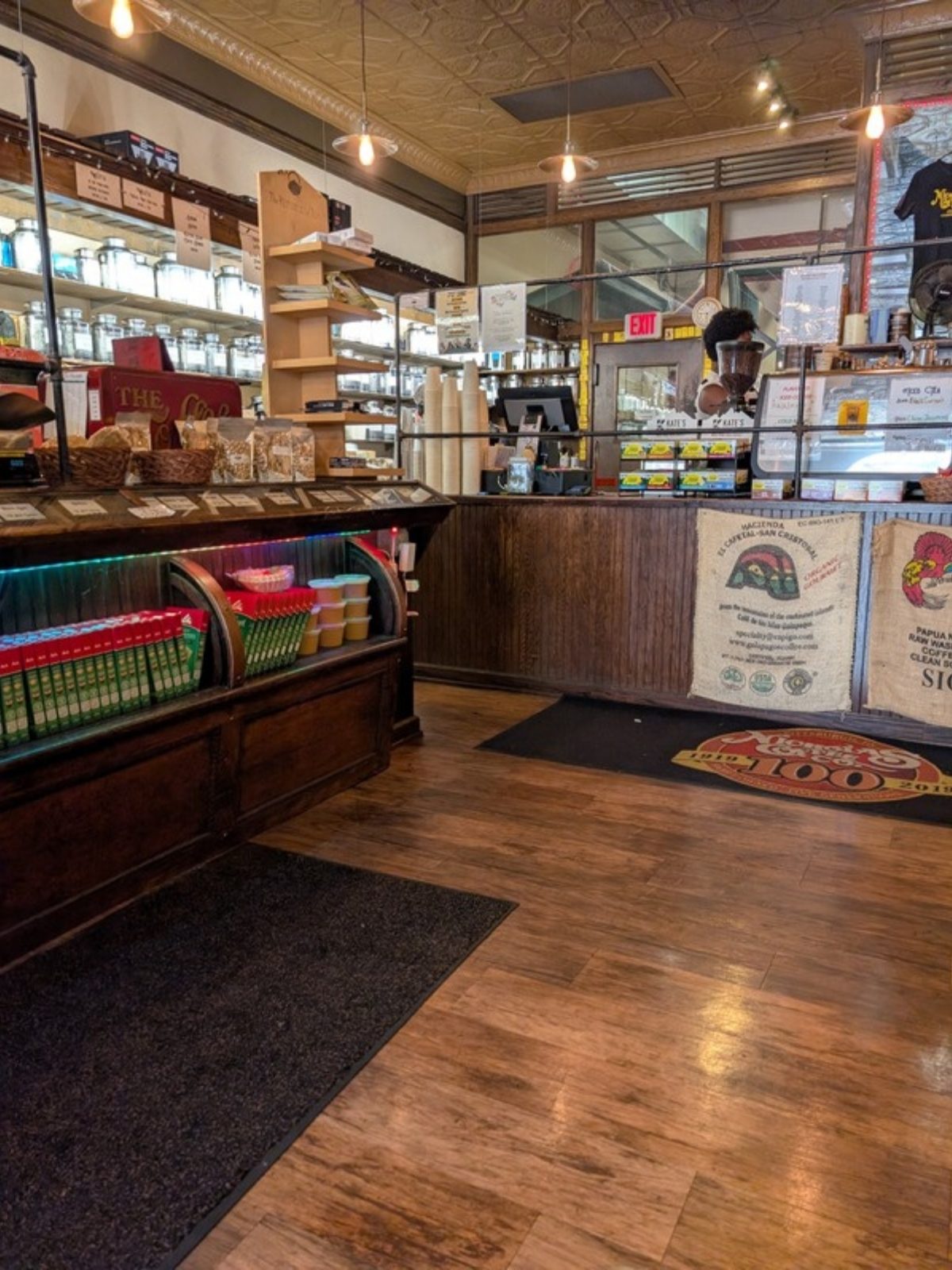 Interior of the shop from seated height. A long dark wooden display case runs down the left side with shelves of glass jars above it. On the right, the service counter is faced with burlap coffee sacks. Between them the wood floor is open and clear, with dark mats laid along it. A pressed tin ceiling runs overhead. One staff member behind the counter has been blurred for privacy.