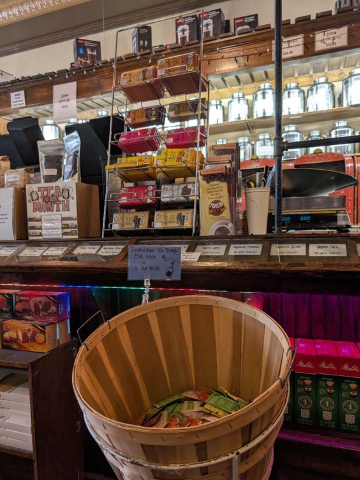 Facing a long wooden display case at seated height. A large wooden bushel basket holding individually wrapped tea bags sits against the front of the case at about lap height, with a small price sign clipped above it. Behind the case, a wire rack of boxed teas and shelves of tall metal tea canisters line the wall.
