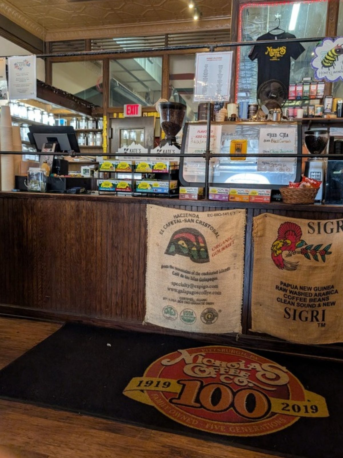 The service counter seen straight on from seated height. The counter front is dark wood hung with two burlap coffee sacks. Above the counter top, a metal rail runs horizontally across the full width at roughly chest height, with a register, a pastry case, handwritten iced tea and cold brew signs, and shelves of syrups and tins behind it. A large round floor mat reading Nicholas Coffee Co. 1919 to 2019 lies on the wood floor in front of the counter.